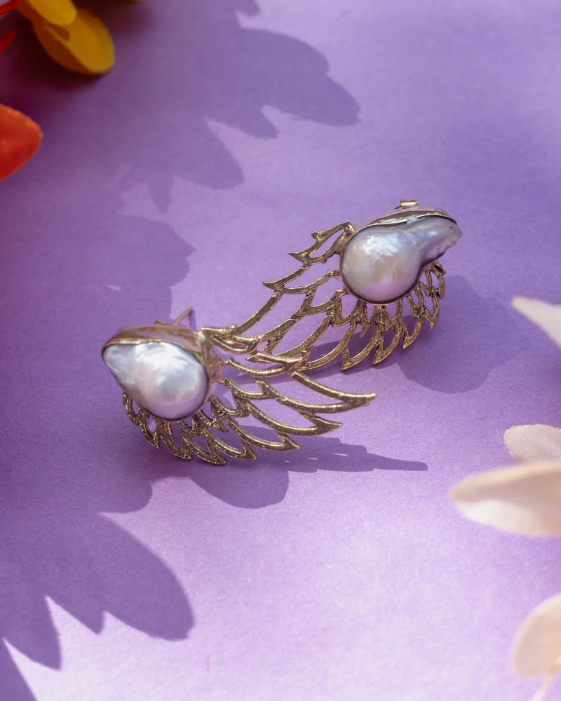 Handmade angel wing pearl earrings photographed on lavender surface with floral accents, highlighting radiant texture and elegant design.