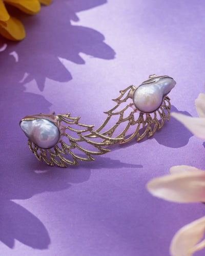 Elegant handmade pearl angel wings studs displayed on purple background with flowers, featuring gold wing frame and lustrous pearls.