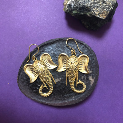 Gold elephant Ganesh earrings placed on black round plate with purple background, highlighting detailed craftsmanship and spiritual design.