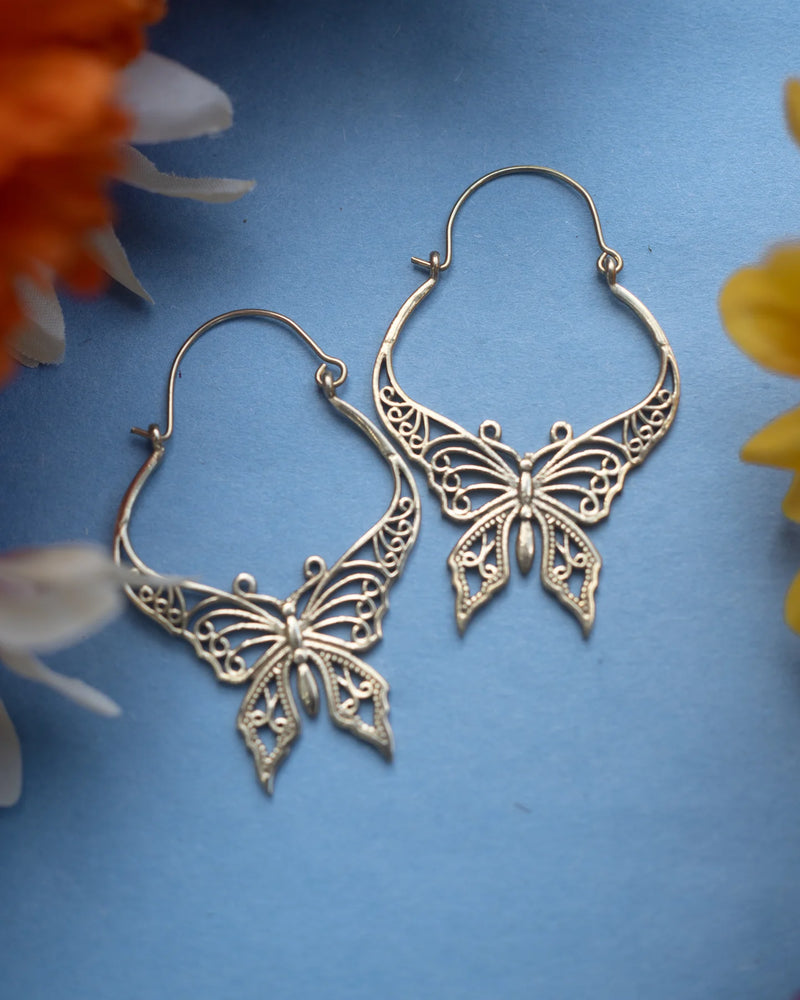 Gold butterfly hoop earrings placed on blue background with floral accents, highlighting delicate filigree craftsmanship and artistic beauty.