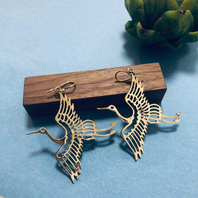 Gold bird-shaped earrings placed on wooden block against blue background, showcasing detailed craftsmanship and artistic style.