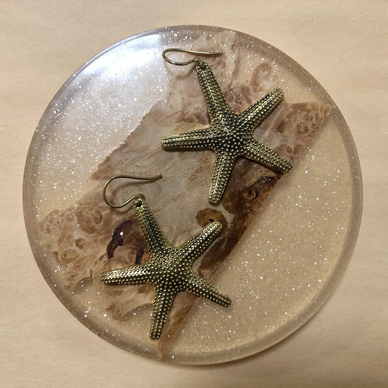 Gold starfish earrings placed on round tray with sand-like texture, showcasing coastal-inspired design and artisanal craftsmanship.