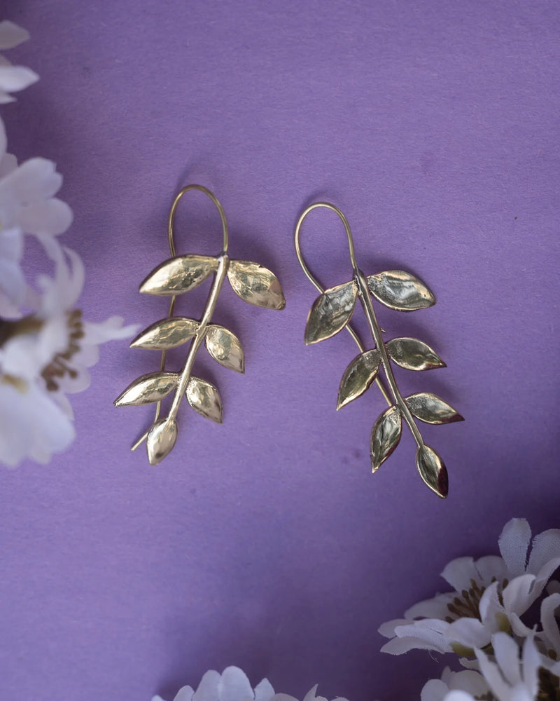 Gold-plated leaf drop earrings placed on purple background with flowers, showcasing nature-inspired elegance and feminine charm.
