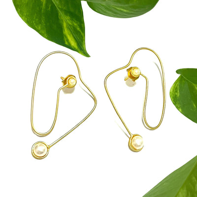Gold abstract wire earrings with white pearls placed on green leaves, highlighting artistic craftsmanship and organic beauty.