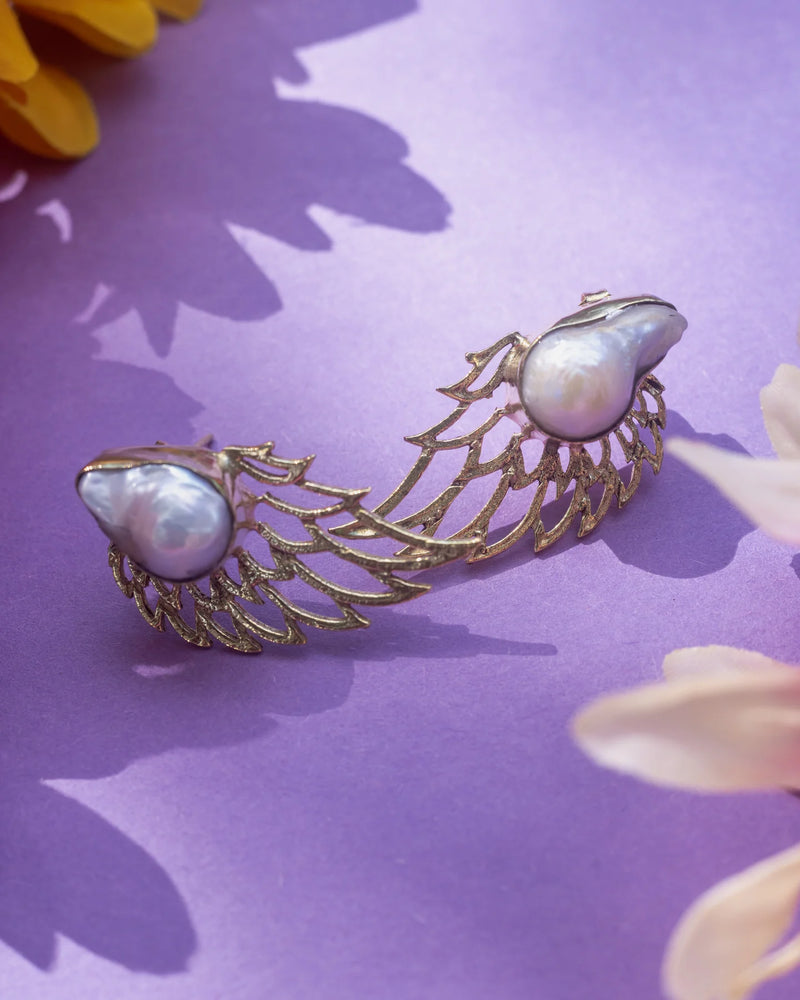 Baroque pearl wing earrings placed on lavender background with flowers, emphasizing artistic composition and radiant pearl sheen.