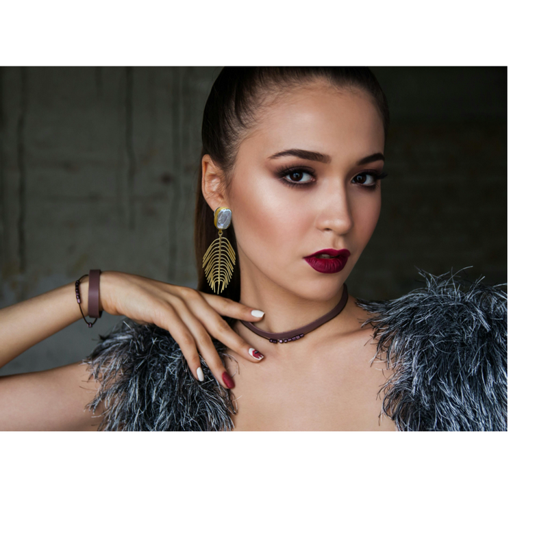 Woman wearing feathered shoulder cover and gold leaf earrings with a dark background