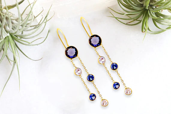 Blue crystal linear drop earrings on white surface with plants, gold-plated elegant design.