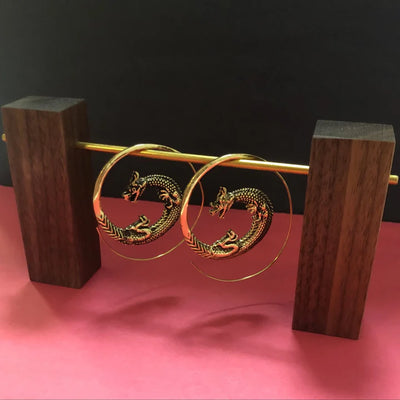Golden dragon hoop earrings displayed on wooden stand with pink background, bold statement jewelry.