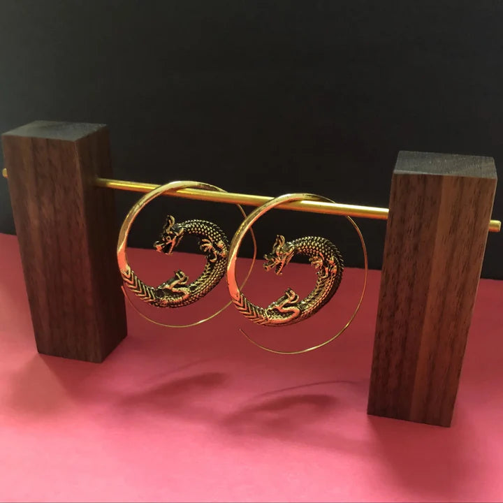 Golden dragon hoop earrings displayed on wooden stand with pink background, bold statement jewelry.