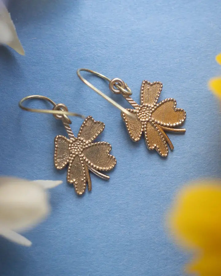 Golden four-leaf clover earrings displayed on blue surface with floral accents, delicate drop design.