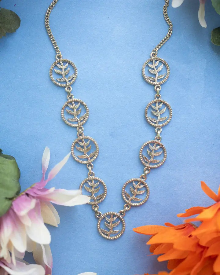 Golden tree of life necklace displayed on blue background with flowers, nature-inspired design.