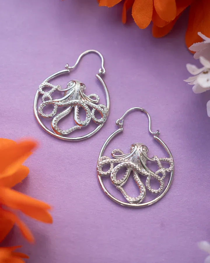 Flatlay of Kraken octopus drop earrings on purple background with floral decoration.