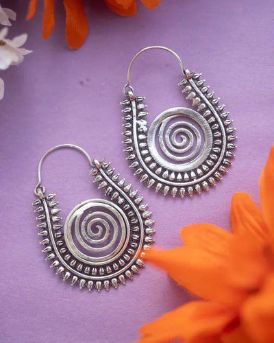 Warli design handcrafted spiral hoop earrings displayed on purple background with flowers.
